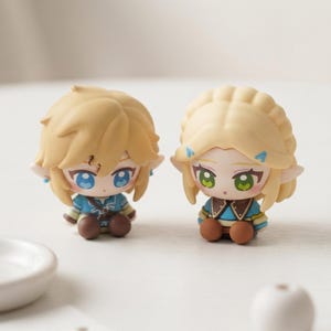 Peut inclure: Deux petites figurines de style chibi avec des cheveux blonds et des oreilles pointues. La figurine de gauche a des yeux bleus et une tunique bleue, tandis que celle de droite a des yeux verts, une coiffure tressée et une tenue bleue et marron.