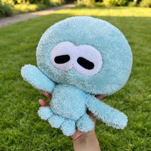 May include: A light blue plush toy shaped like a squid with a round head and textured fabric. It features two large white oval eyes with black pupils. The plush has several small tentacles at the base and two extended arms on the sides.