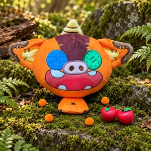 May include: A plush toy shaped like a creature with orange fur, a dark brown patch on its head, and a single yellow horn. It features a button eye in blue and one in green, a pig-like snout, and two small red apple figurines nearby.