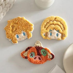 Puede incluir: Tres parches textiles con acabado de tufting que representan cabezas de personajes estilizadas. Dos parches muestran figuras con cabello amarillo y ojos azules, mientras que el tercero muestra una criatura marrón y naranja con un cuerno.