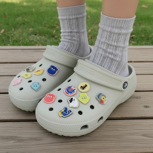 May include: A pair of light gray slip-on clogs with a textured toe cap and heel strap, adorned with various colorful, small, flat character-themed charms inserted into the ventilation holes, worn with light gray ribbed knit crew socks.