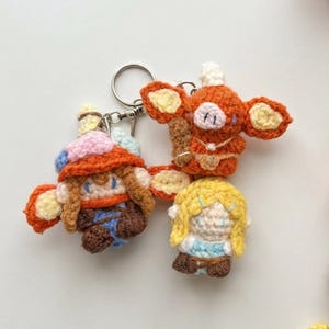May include: Three small crocheted figures attached to a silver metal keychain. One figure is a brown-haired character in a colorful hat, another is an orange creature with large ears, and the third is a blonde character in a light blue outfit.