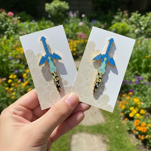 May include: Two enamel pins shaped like swords with blue, light blue, and black coloring. The sword design features a winged hilt and a blade that transitions from a solid color to a dark, swirling pattern at the tip, mounted on white cards.