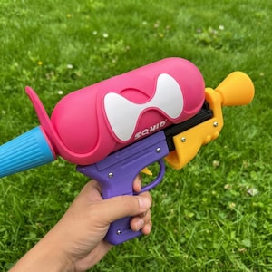 May include: A plastic water blaster toy featuring a bright pink cylindrical tank with a white bow-shaped graphic and the word SQUID printed on the side. The toy has a purple handle and trigger, a blue nozzle, and a yellow rear section.