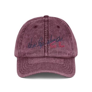 May include: A burgundy acid-wash baseball cap with a six-panel crown and a curved brim. The front features embroidered text in dark blue and pink thread that reads Never Stop... Wear Out... For Them, centred above the brim on the front panels.