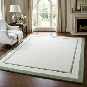 May include: A rectangular area rug featuring a large, solid off-white center panel framed by a wide, sage green border. The rug has a clean, flat-woven appearance with a distinct rectangular outline separating the inner and outer sections.