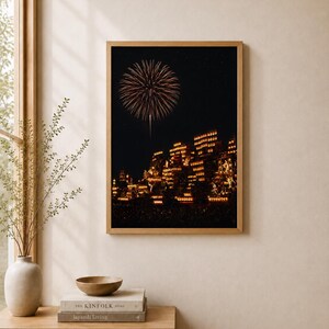 May include: A rectangular wall art print in a light wood frame featuring a night scene of a large firework burst above a cluster of illuminated buildings with warm glowing lights against a dark sky, displayed on a neutral wall.