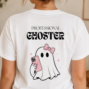 Puede incluir: Una camiseta blanca de manga corta con un gráfico en la espalda. El texto dice PROFESSIONAL GHOSTER en negro. Debajo del texto hay una ilustración de un fantasma blanco con un lazo rosa, sosteniendo un teléfono inteligente rosa.