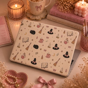 May include: A laptop skin featuring a cream-colored background with a repeating pattern of black cats, witch hats with pink bows, brown broomsticks, pink potion bottles, open spell books, lit candles, crescent moons, and small pink bows.