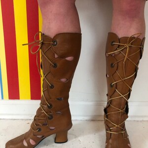 May include: Brown leather boots with cutouts and lace-up detailing. The boots have a block heel and a pointed toe.