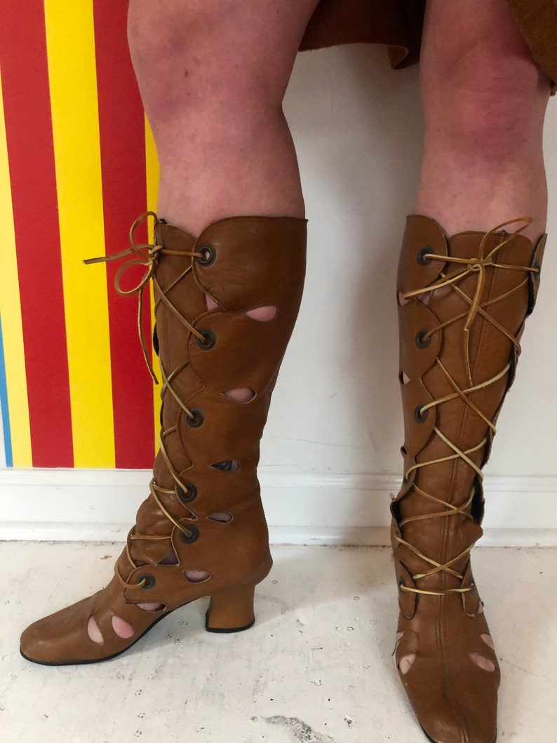 May include: Brown leather boots with cutouts and lace-up detailing. The boots have a block heel and a pointed toe.