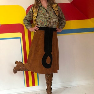 May include: A brown suede skirt with a black velvet waistband and a black suede detail. The skirt is knee-length and has a flared silhouette. The person wearing the skirt is also wearing a green paisley blouse with a gold vest and brown boots.