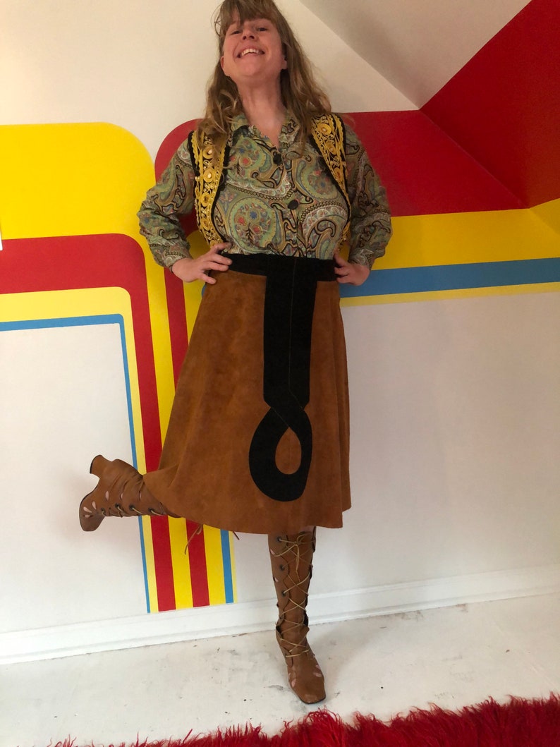 May include: A brown suede skirt with a black velvet waistband and a black suede detail. The skirt is knee-length and has a flared silhouette. The person wearing the skirt is also wearing a green paisley blouse with a gold vest and brown boots.