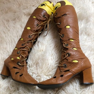 May include: A pair of brown leather knee-high boots with lace-up fronts and chunky heels. The boots have cutouts and yellow accents.