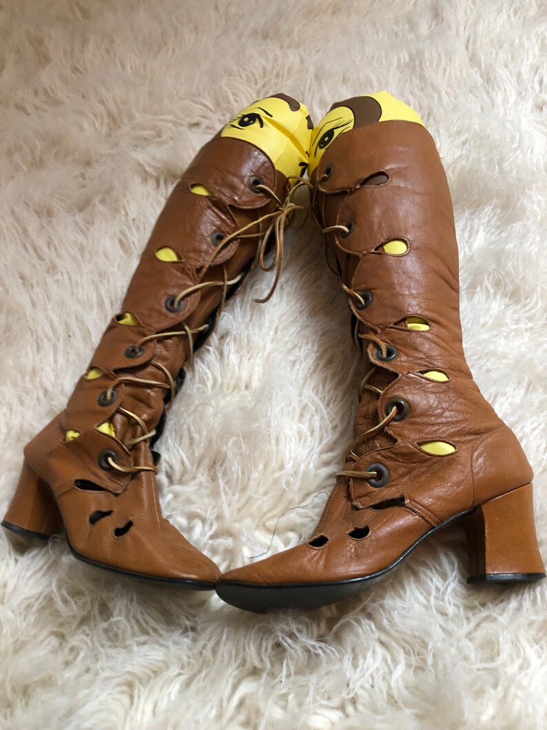 May include: A pair of brown leather knee-high boots with lace-up fronts and chunky heels. The boots have cutouts and yellow accents.