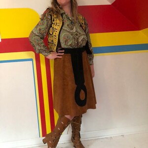 May include: A woman wearing a brown suede skirt with a black belt and a paisley print blouse with a gold embroidered vest. She is wearing brown leather boots with cutouts and laces.