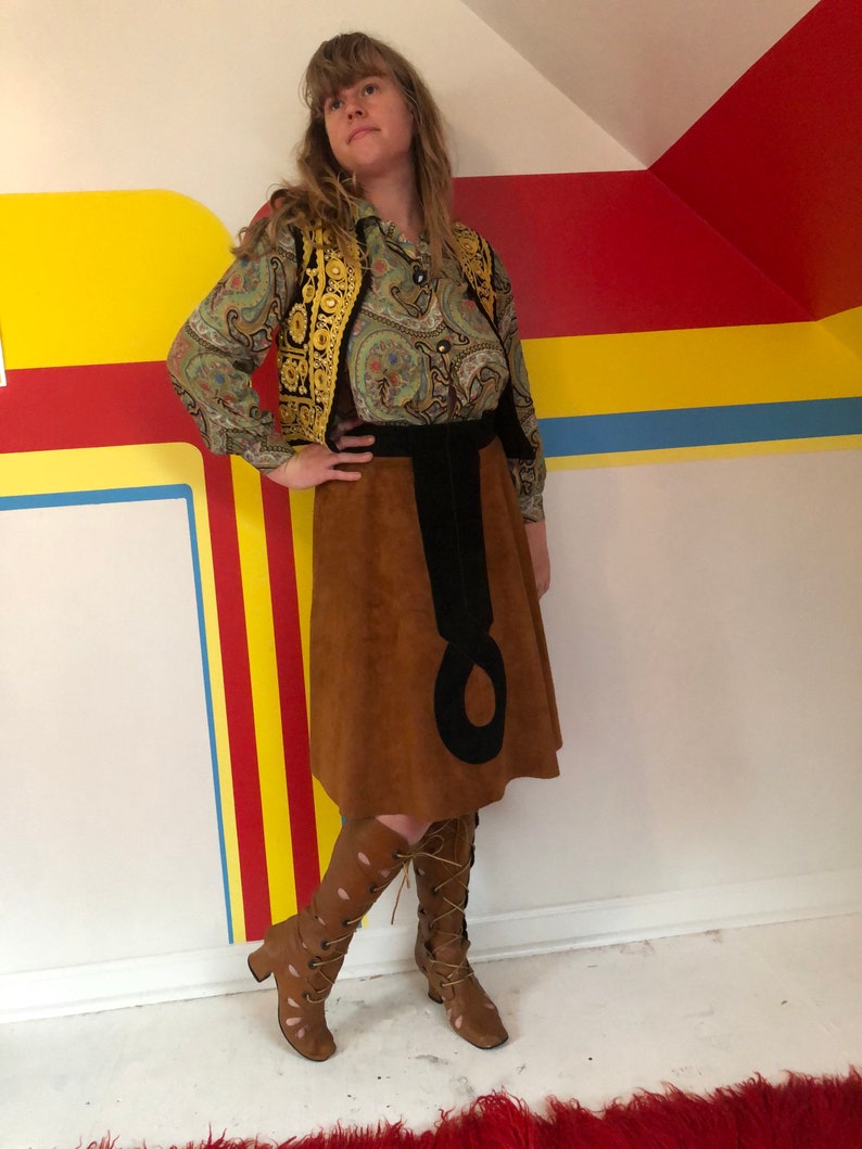 May include: A woman wearing a brown suede skirt with a black belt and a paisley print blouse with a gold embroidered vest. She is wearing brown leather boots with cutouts and laces.