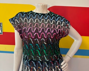 May include: A black sequined top with a wavy pattern of colorful sequins. The top has short sleeves and is displayed on a mannequin. The bottom of the mannequin is wearing a blue fringe skirt. The background is a colorful wall.