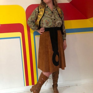 May include: A brown suede skirt with a black design, paired with a green paisley blouse and a gold embroidered vest. The outfit is completed with brown lace-up boots.