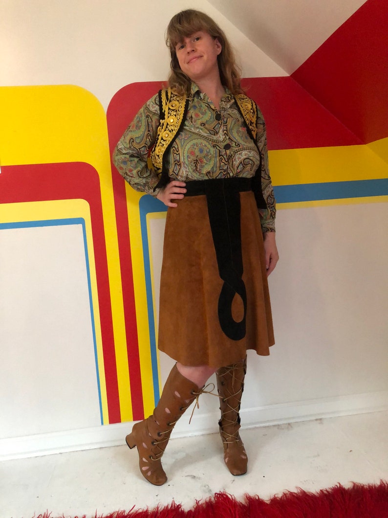 May include: A brown suede skirt with a black design, paired with a green paisley blouse and a gold embroidered vest. The outfit is completed with brown lace-up boots.