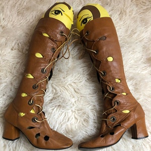 May include: A pair of brown leather knee-high boots with lace-up fronts and cut-out details. The boots have a block heel and a pointed toe.