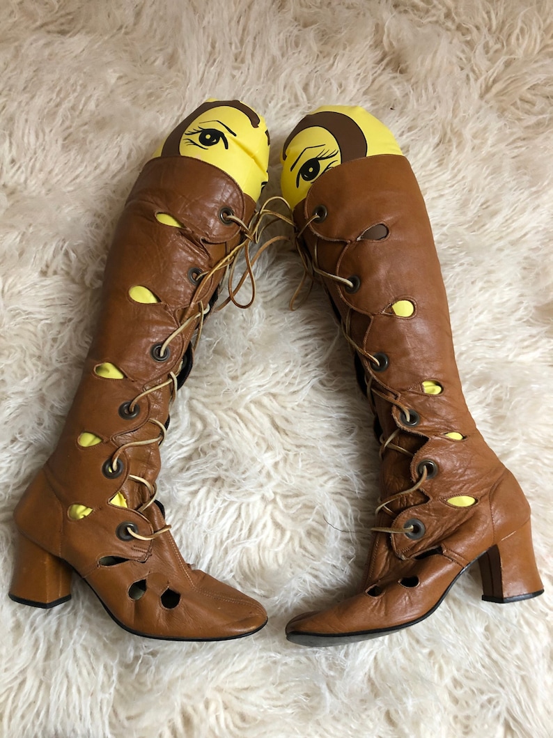 May include: A pair of brown leather knee-high boots with lace-up fronts and cut-out details. The boots have a block heel and a pointed toe.