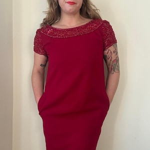Peut inclure: Une robe rouge avec un col orné de perles et des manches courtes. La robe a un design simple avec des poches. La robe arrive au genou et est faite d'un tissu lisse. La robe est d'un rouge vif.