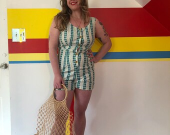 May include: A woman wearing a white and green striped button-up jumpsuit with a white background and green and blue geometric pattern. She is holding a beige mesh bag with a wooden handle. She is standing in front of a wall with yellow, red, and blue stripes.
