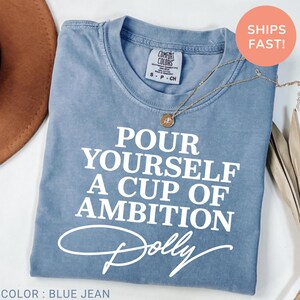 May include: A blue jean colored t-shirt with white text that reads "POUR YOURSELF A CUP OF AMBITION" followed by the name "Dolly" in a script font. The shirt features a crew neckline and a Comfort Colors brand label on the inside collar.