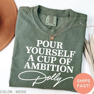 May include: A moss green short-sleeve t-shirt featuring the white text "POUR YOURSELF A CUP OF AMBITION" followed by the signature "Dolly" in white script. The shirt has a crew neckline and a Comfort Colors brand label at the inner collar.