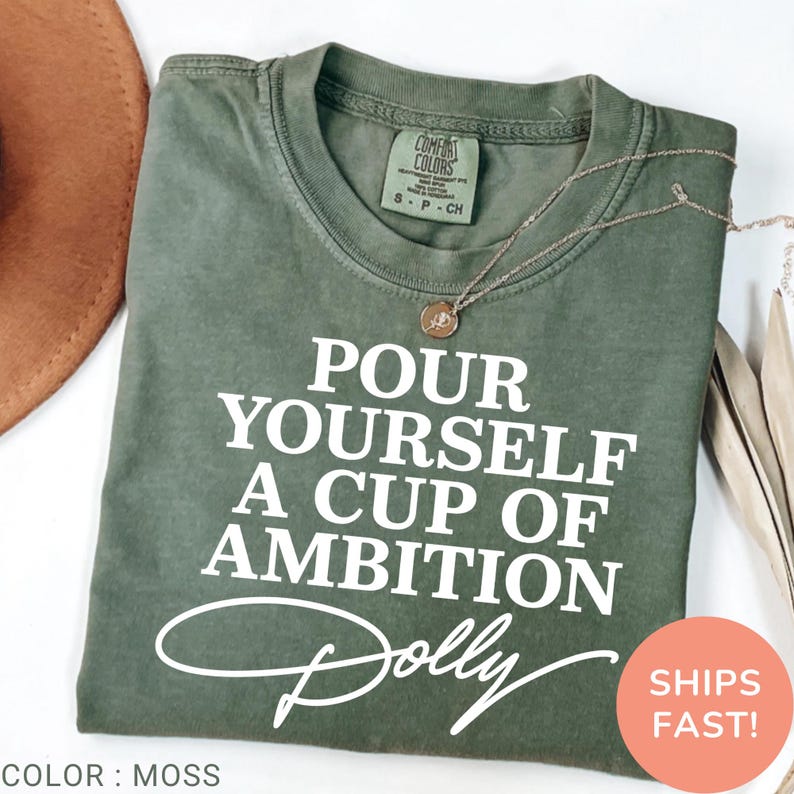 May include: A moss green short-sleeve t-shirt featuring the white text "POUR YOURSELF A CUP OF AMBITION" followed by the signature "Dolly" in white script. The shirt has a crew neckline and a Comfort Colors brand label at the inner collar.