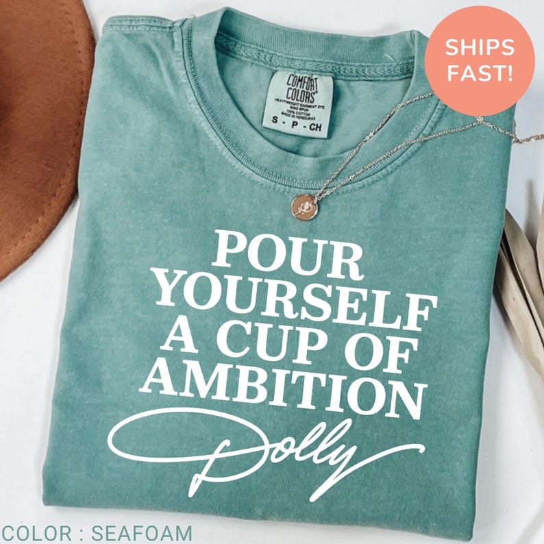 May include: A seafoam green short-sleeve t-shirt with white text that reads "POUR YOURSELF A CUP OF AMBITION" followed by the name "Dolly" in a script font. The shirt features a crew neckline and a Comfort Colors brand label at the collar.