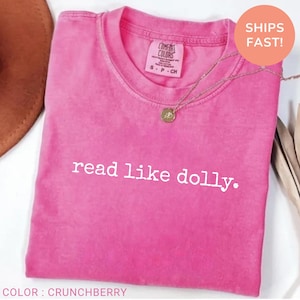 May include: A bright pink short-sleeved t-shirt with a crew neckline. The shirt features the white text read like dolly. printed across the chest. A gold-toned necklace with a circular pendant rests on the fabric near the collar area.