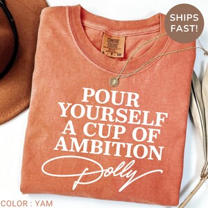 May include: A short-sleeved crew neck t-shirt in a washed-out terracotta orange color. The shirt features white screen-printed text that reads, "POUR YOURSELF A CUP OF AMBITION" with the name "Dolly" written in a cursive script font underneath.
