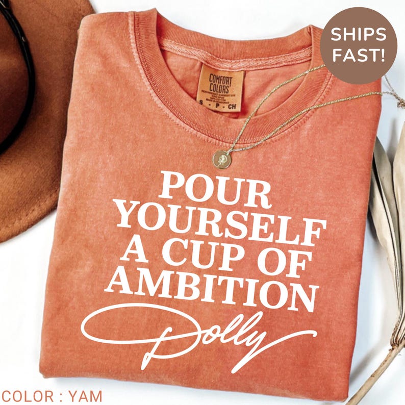 May include: A short-sleeved crew neck t-shirt in a washed-out terracotta orange color. The shirt features white screen-printed text that reads, "POUR YOURSELF A CUP OF AMBITION" with the name "Dolly" written in a cursive script font underneath.