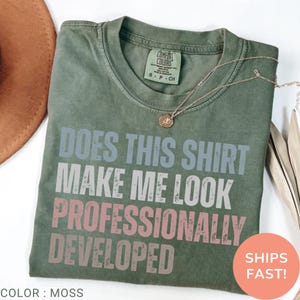 Peut inclure: Un t-shirt à manches courtes vert mousse avec le texte "DOES THIS SHIRT MAKE ME LOOK PROFESSIONALLY DEVELOPED" dans une police usée. Le t-shirt présente une étiquette Comfort Colors au col et est accompagné d'un collier doré délicat.