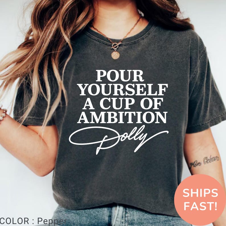 May include: A charcoal gray short-sleeve t-shirt featuring the white text "POUR YOURSELF A CUP OF AMBITION" followed by the signature "Dolly" in a cursive script. The shirt has a crew neckline and a vintage-style washed fabric finish.