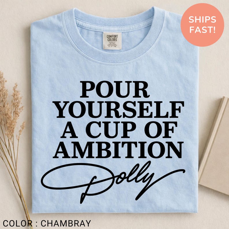 May include: A light blue crew neck t-shirt featuring the black text "POUR YOURSELF A CUP OF AMBITION" followed by the word "Dolly" in a cursive script font. The shirt has a Comfort Colors brand label on the inside of the collar.