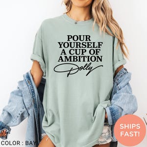May include: A sage green short-sleeve t-shirt featuring black text that reads POUR YOURSELF A CUP OF AMBITION with the name Dolly written in a cursive script underneath. The shirt has a crew neckline and is worn with a blue denim jacket.