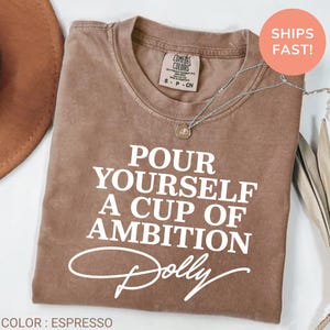 May include: An espresso-colored short-sleeve t-shirt with a crew neckline. The shirt features white text that reads, "POUR YOURSELF A CUP OF AMBITION" with the name "Dolly" written in a cursive script font underneath the main text.