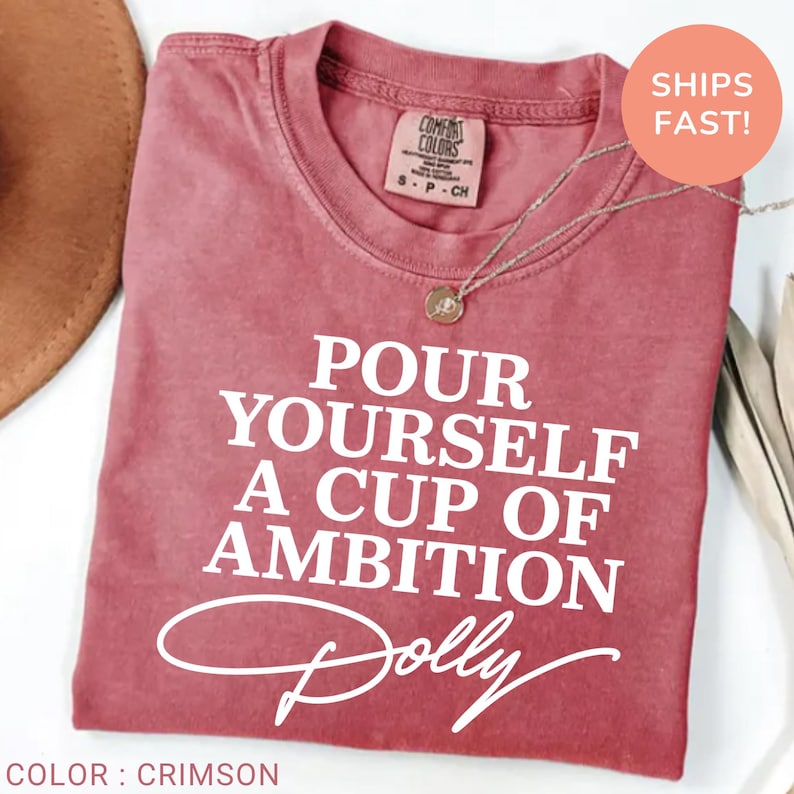 May include: A crimson short-sleeved t-shirt featuring the white text "POUR YOURSELF A CUP OF AMBITION" followed by the signature "Dolly" in a script font. The shirt has a crew neckline and a Comfort Colors brand label at the inner collar.