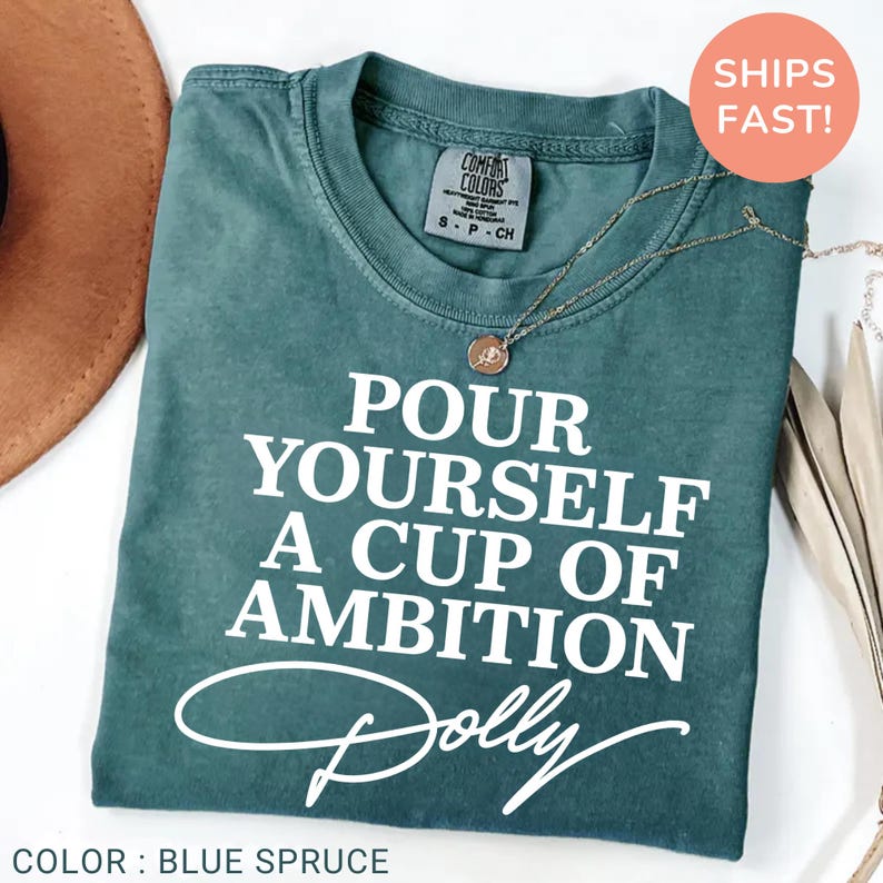 May include: A blue spruce short-sleeve t-shirt featuring the white text "POUR YOURSELF A CUP OF AMBITION" followed by the word "Dolly" in a cursive script. The shirt has a crew neckline and a Comfort Colors brand label at the inner collar.