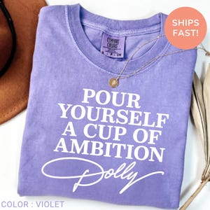 May include: A violet short-sleeved t-shirt with a crew neckline featuring the white printed text "POUR YOURSELF A CUP OF AMBITION" followed by the word "Dolly" in a cursive script font. A thin gold-toned necklace rests on the fabric.