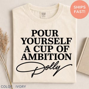 May include: An ivory short-sleeved t-shirt featuring the black printed text "POUR YOURSELF A CUP OF AMBITION" followed by the word "Dolly" in a cursive script font. The shirt has a crew neckline and a Comfort Colors brand label at the neck.