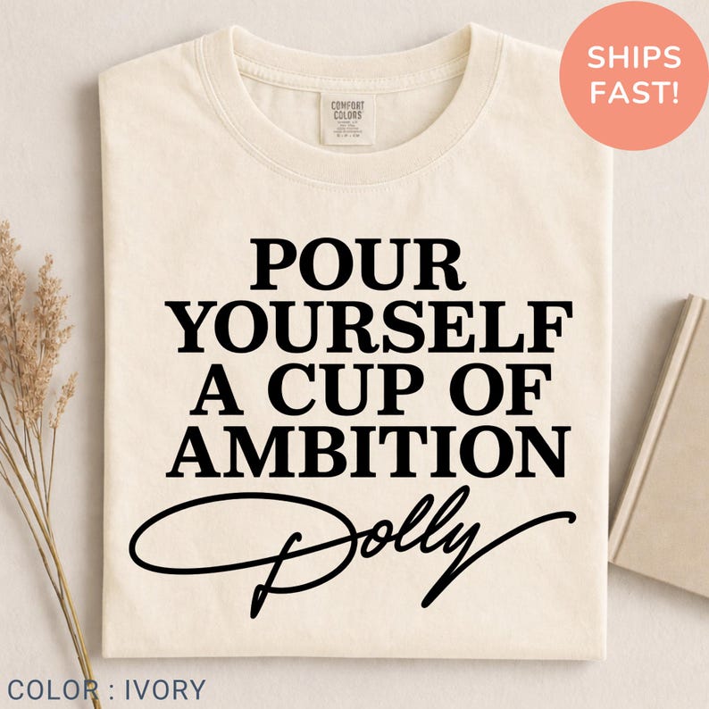 May include: An ivory short-sleeved t-shirt featuring the black printed text "POUR YOURSELF A CUP OF AMBITION" followed by the word "Dolly" in a cursive script font. The shirt has a crew neckline and a Comfort Colors brand label at the neck.