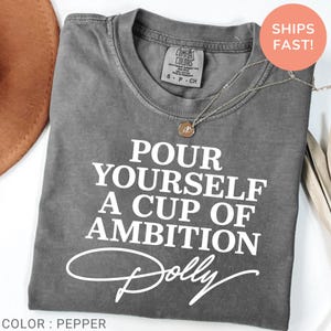 May include: A pepper-colored short-sleeve t-shirt with white text that reads POUR YOURSELF A CUP OF AMBITION and the name Dolly in a script font. The shirt features a crew neckline and a Comfort Colors brand label on the inside collar.