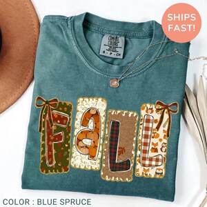 May include: A blue spruce t-shirt featuring the word FALL in large block letters. Each letter has a unique pattern: F with leaves, A with pumpkins, L with red and black plaid, and L with foxes. The letters are decorated with bows.