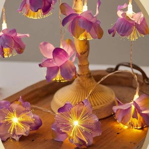 May include: A string of decorative lights featuring purple fabric flower petals. Each flower contains a central cluster of thin, glowing yellow fiber optic filaments that emit a bright light. The lights are connected by a thin, clear wire.