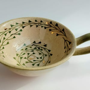 Puede incluir: Una taza de cerámica color crema con un cuenco ancho y poco profundo y una sola asa. El interior presenta un patrón de enredadera verde pintado a mano con pequeñas hojas en forma de corazón y detalles en oro. El asa tiene un degradado verde.