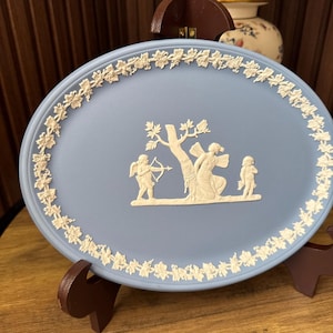 Może przedstawiać: Owalna, jasnoniebieska taca ceramiczna z białymi zdobieniami w reliefie. Krawędź przedstawia ciągły motyw winorośli i winogron. Środek ukazuje klasyczną scenę ze skrzydlatą postacią celującą z łuku do drzewa, siedzącą postacią oraz małym dzieckiem.